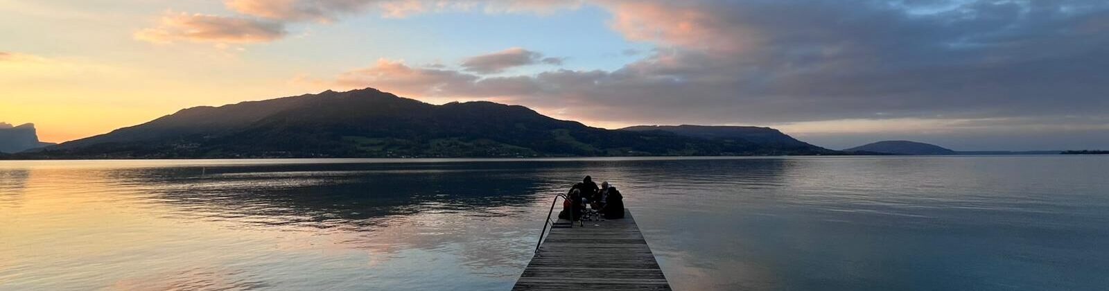 Holzsteg, der über einen ruhigen See führt, mit zwei Personen am Ende, während die Sonne untergeht.