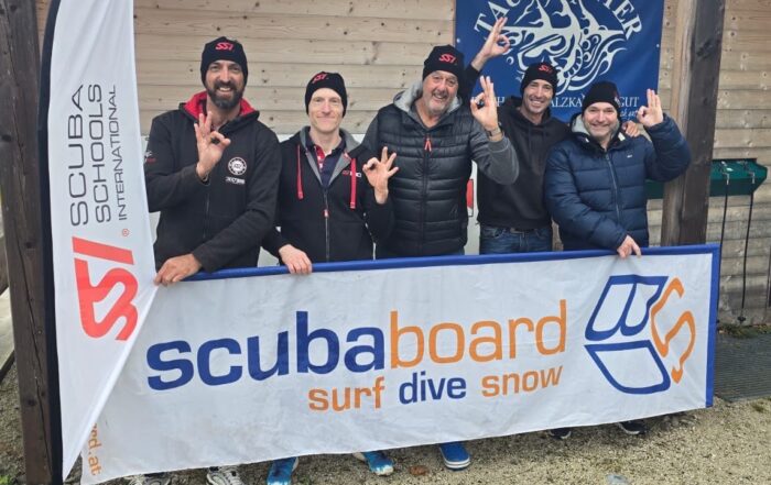Fünf Taucher stehen vor einem Banner von Scubaboard und zeigen das Handzeichen für Tauchen.