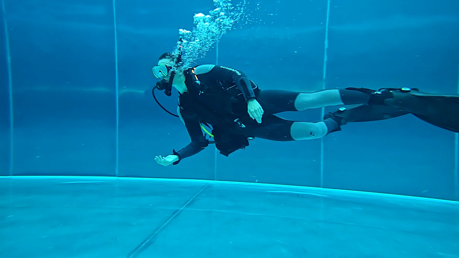 Open Water Diver im Pooltraining mit Scubapro-Ausrüstung Ein Taucher in schwarzem Neoprenanzug schwebt im blauen Wasser und atmet aus der Tauchflasche auf.