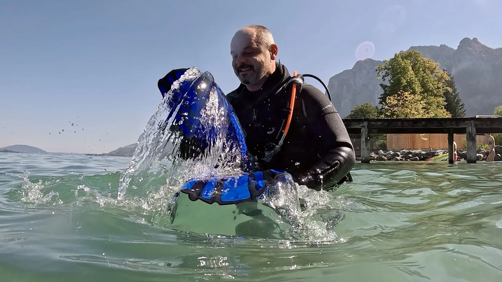GPTempDownload-10 Ein Taucher im schwarzen Neoprenanzug hält blaue Ausrüstung aus dem Wasser, während Spritzwasser hoch aufsteigt.