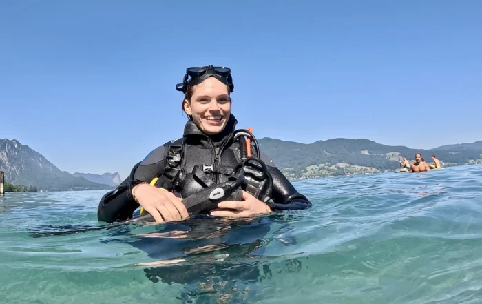 Eine lächelnde Taucherin im schwarzen Neoprenanzug hält sich im klaren Seewasser über Wasser.