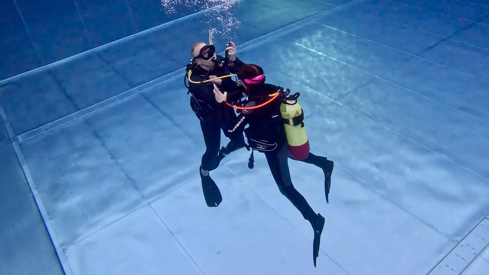 GPTempDownload-2 Zwei Taucher üben im Pool eine Unterwasserübung, einer mit gelber Tauchflasche und pinker Maske.