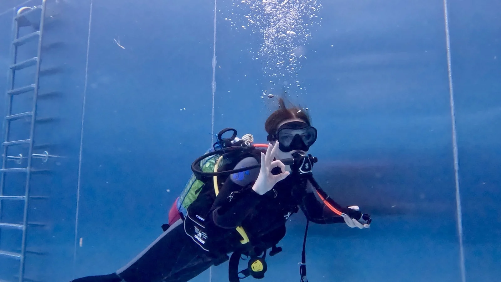 GPTempDownload-5 Eine Taucherin schwebt im Pool unter Wasser und zeigt das OK-Zeichen, während Luftblasen nach oben steigen.
