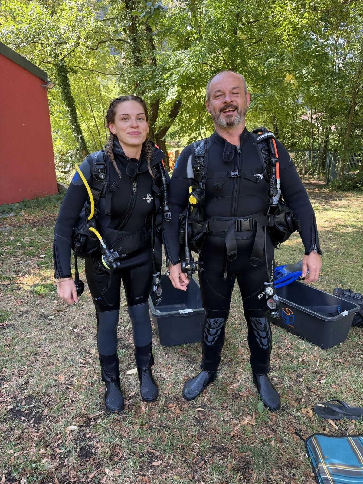 IMG_2512 Ronja und Mario stehen im schwarzen Tauchanzug mit Tauchflaschen und Ausrüstung im Grünen beim Open Water Diver Kurs.