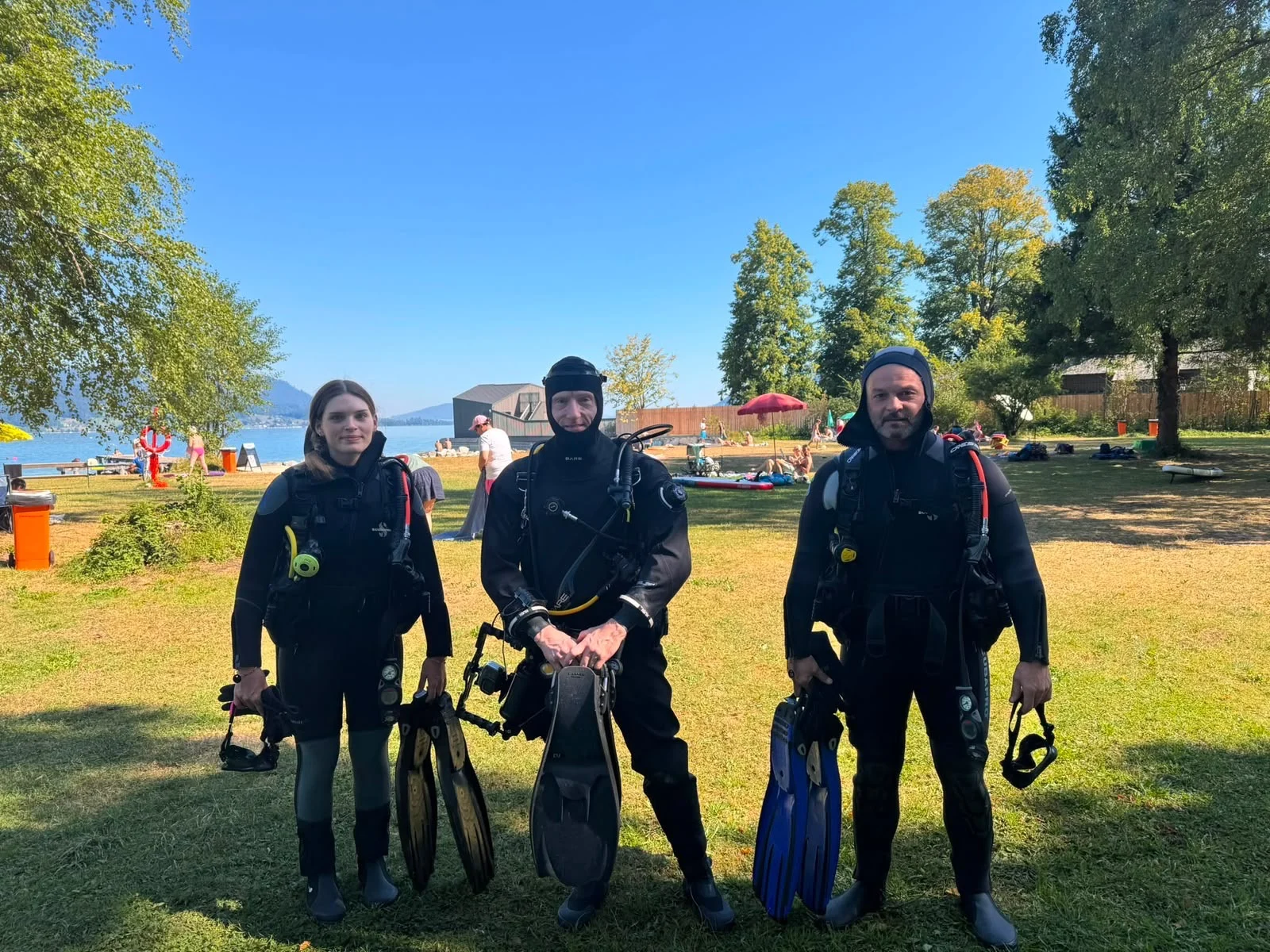 e97a1c44-9e58-4e2c-aaa6-e7d3d6481a84 Ronja und Mario stehen mit ihrem Tauchlehrer im schwarzen Tauchanzug am Ufer des Attersees nach dem Open Water Diver Kurs.